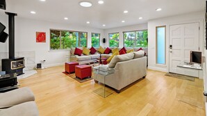 A modern living room with a sofa, red ottoman, and glass coffee table. There is a fireplace, large windows, and a door with a glass panel.