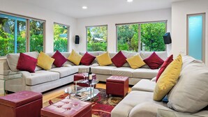 A modern living room with a sectional sofa, red and yellow cushions, a glass coffee table, and large windows offering a view of greenery outside.