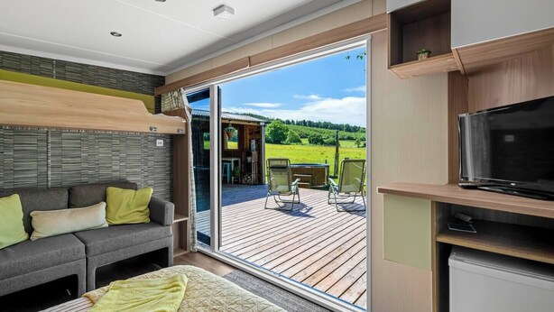 Interior moderno de una caravana con sofá, cama, televisor y puerta corrediza de vidrio que da a una terraza exterior.
