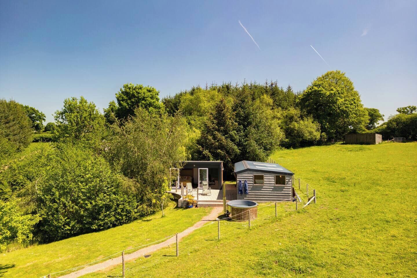 Eine kleine Holzhütte mit einer Terrasse und einem Whirlpool, umgeben von üppigem Grün und Bäumen.