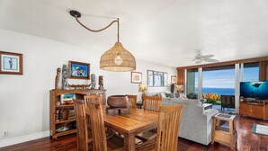 A spacious living room with a wooden dining table, chairs, and a large TV. There are framed pictures on the walls and a ceiling fan. - KKSRC#1-101 Direct Oceanfront, Best North Ground Floor Corner, Remodeled, AC! (KAILUA KONA)