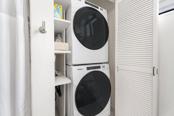 Ett tvättrum med två tvättmaskiner och torktumlare från Whirlpool staplade ovanpå varandra, en hylla med tvättmedel och en vit dörr med ett handtag.