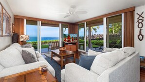 A spacious living room with a large sliding glass door leading to a balcony with a view of the ocean. There is a ceiling fan, a coffee table, and a TV on a wooden stand. - KKSRC#1-101 Direct Oceanfront, Best North Ground Floor Corner, Remodeled, AC! (KAILUA KONA)