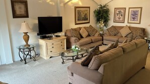 A living room with a beige sofa, a glass coffee table, a TV on a wooden cabinet, and a lamp on a side table. - Charming 4-bedroom villa with WiFi, AC in wonderful Kissimmee (Kissimmee)