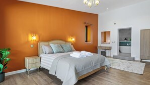 Ein Schlafzimmer mit Bett, Nachttischen, einer Stehlampe, einer Topfpflanze, einem Kronleuchter und Blick ins Bad.