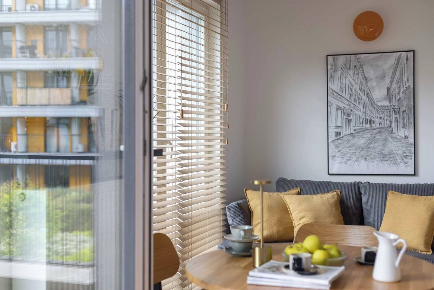 Una habitación con vistas a un edificio, una mesa con frutas y una jarra, un sofá con cojines y una obra de arte enmarcada en la pared.