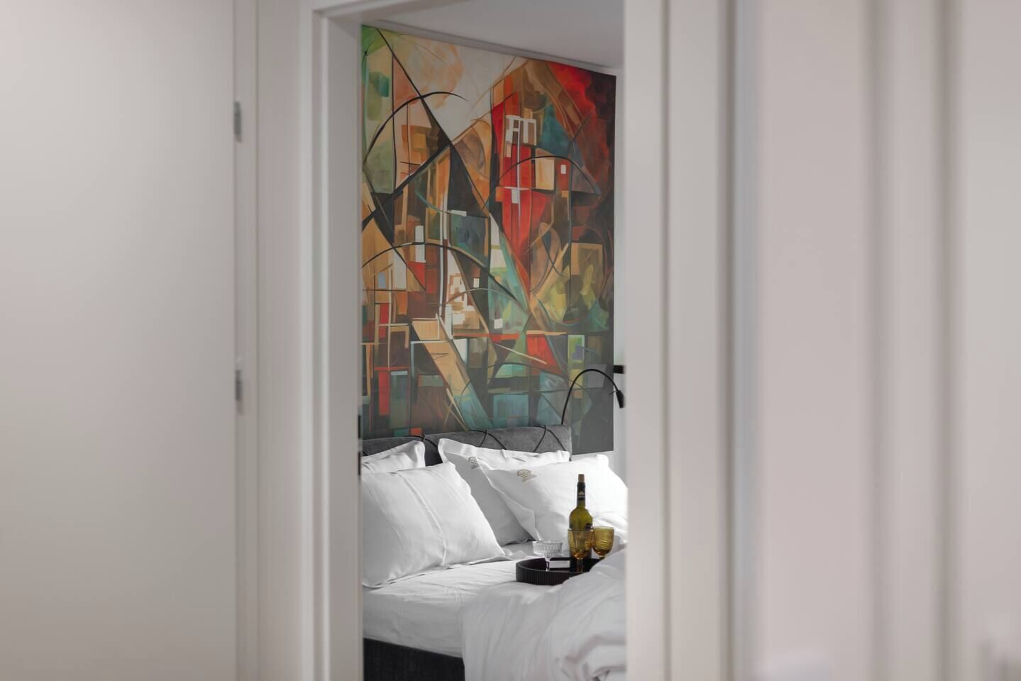 Quarto com cama, roupa de cama branca, bandeja com copos e garrafa, e uma pintura abstrata colorida na parede.