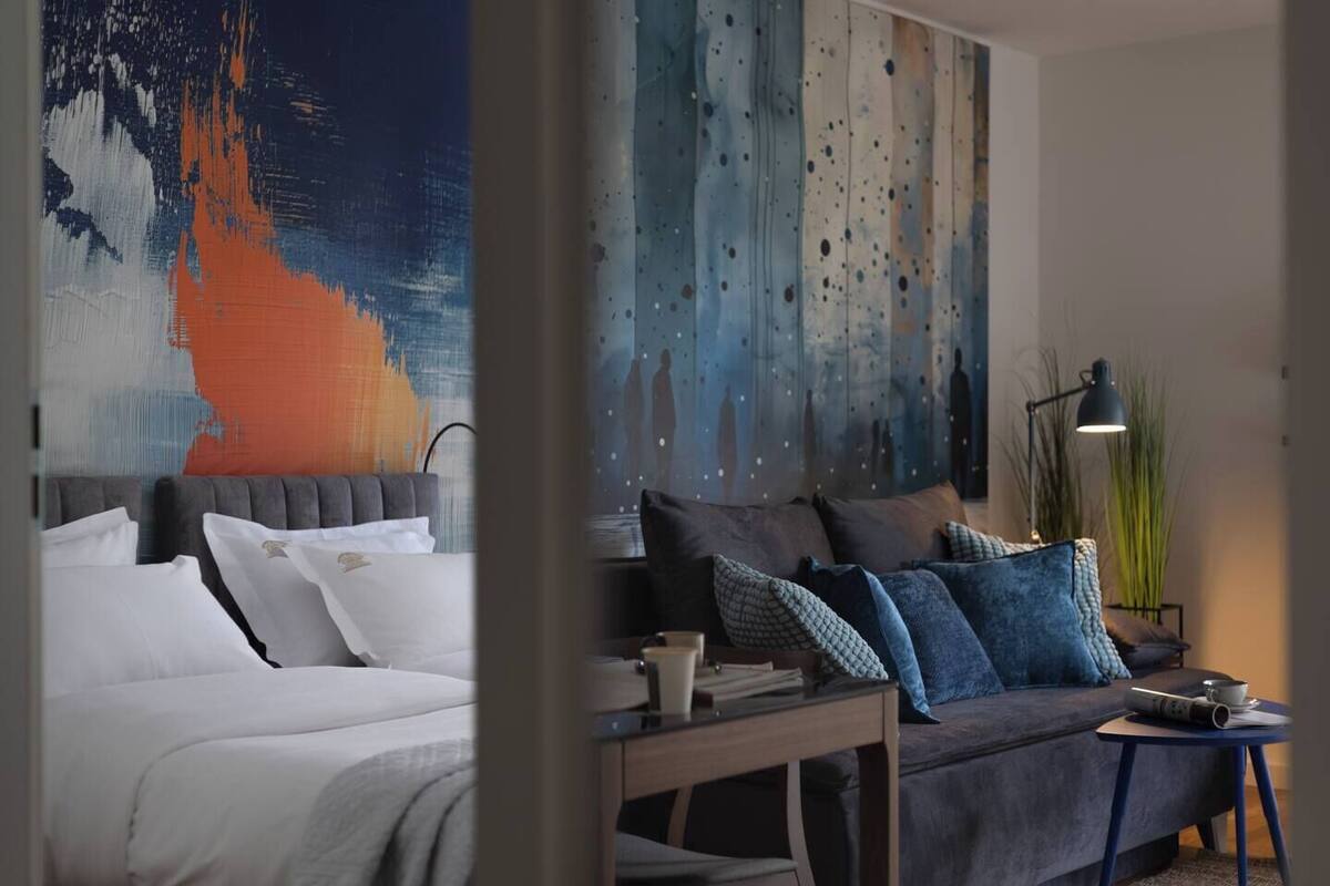 A modern hotel room with a bed, a sofa, a desk, and abstract wall art.