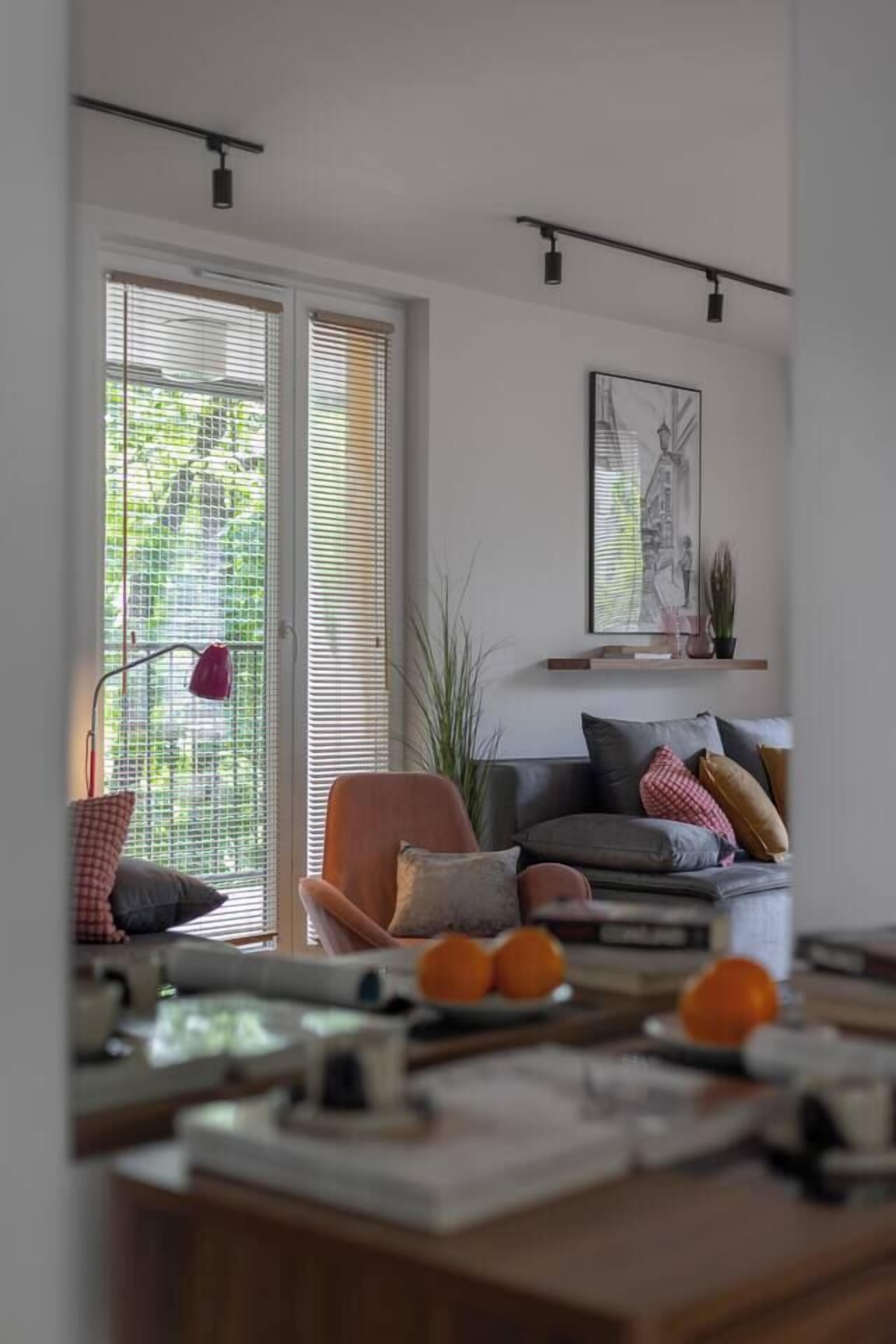 A modern living room with a sofa, a coffee table, and a desk. There are two windows with blinds, a red lamp, and a framed picture on the wall.