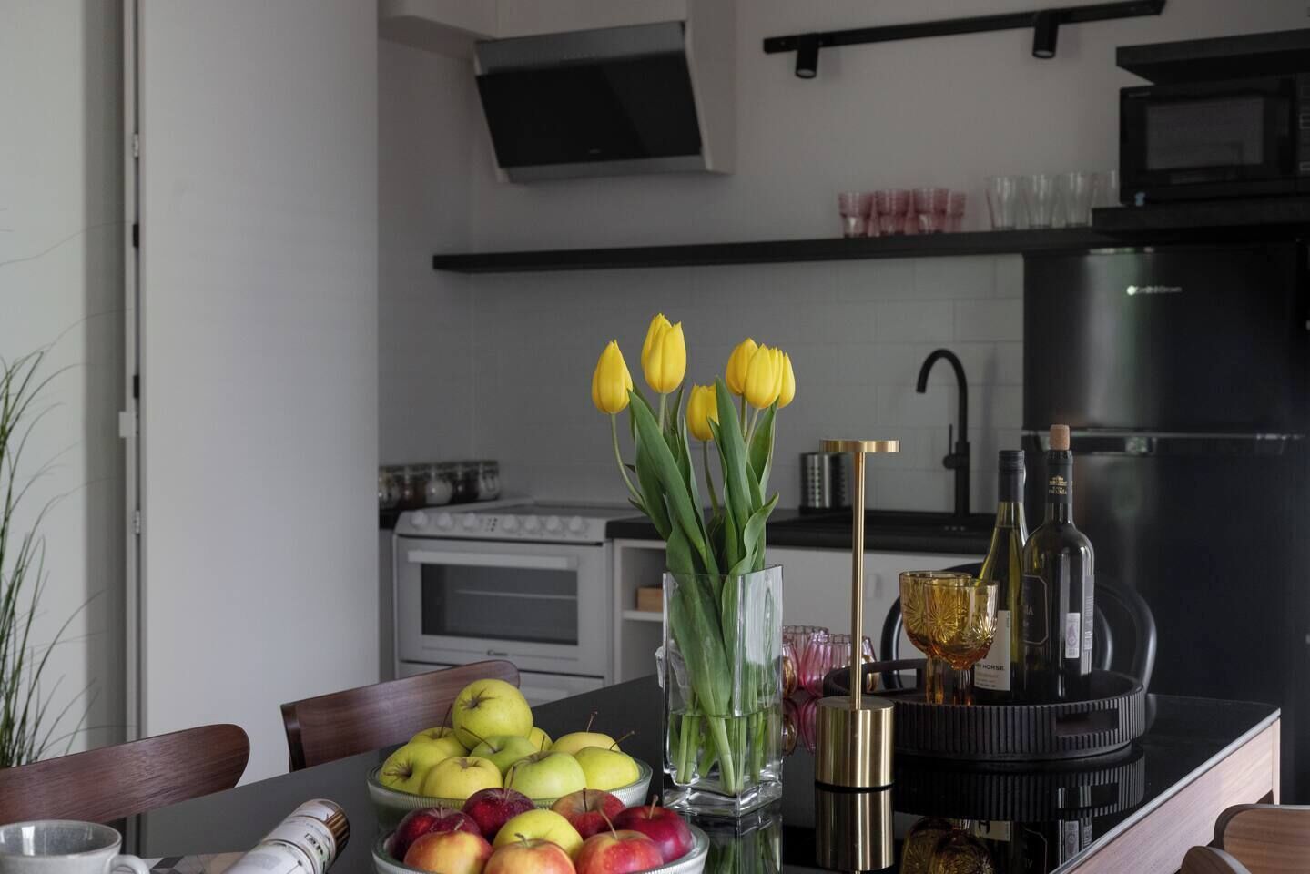 Uma cozinha moderna com fogão, forno e geladeira preta. Há uma mesa de jantar com uma tigela de maçãs e um vaso de tulipas amarelas.