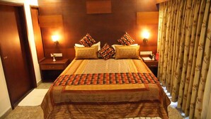 Une chambre d’hôtel avec un grand lit, deux tables de chevet, une petite table, une tête de lit en bois et un rideau.