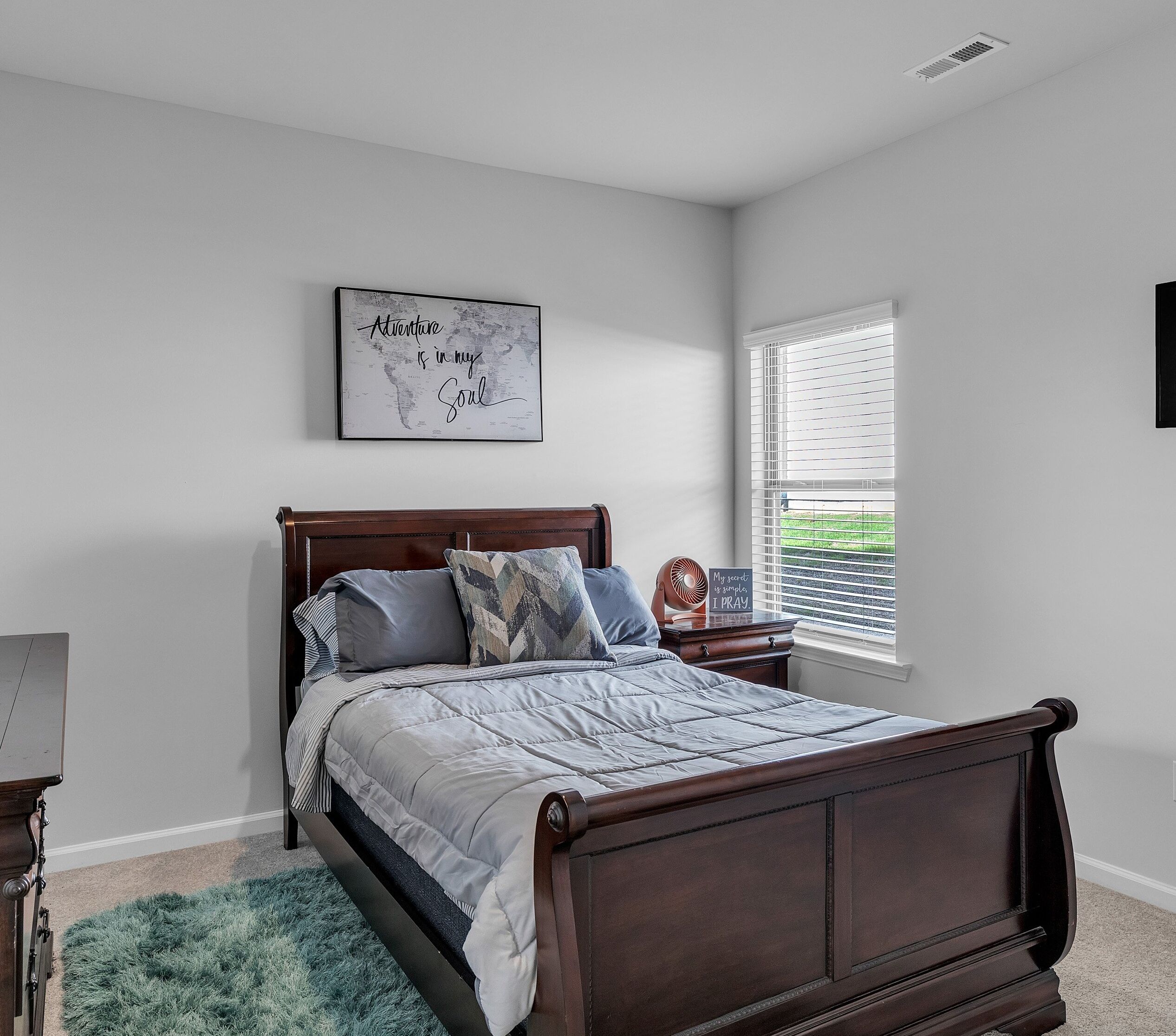 A bedroom with a wooden bed, a dresser, a mirror, a window with blinds, and a wall-mounted shelf.