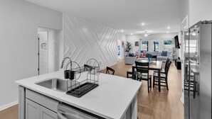 Une cuisine moderne avec un plan de travail blanc, des appareils en acier inoxydable et un coin repas comprenant un canapé gris et une table en bois.