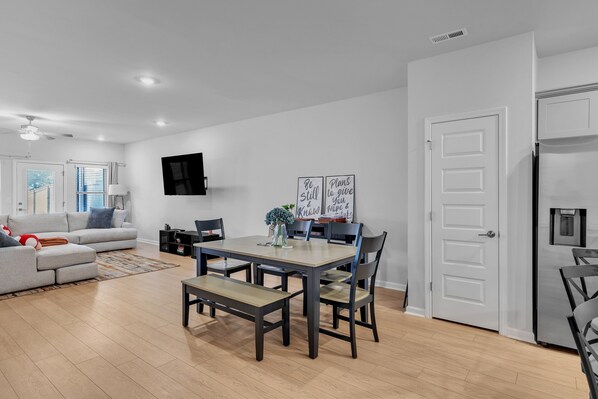 A modern living room with a grey sofa, a dining table with chairs, a flat-screen TV mounted on the wall, and a door leading to another room. - Modern Nest - Huntsville Hideaway 3 Bedroom Home (Huntsville)