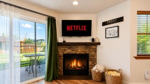A living room with a stone fireplace, a TV mounted above it, and a view of the outdoors. - Edelweiss Escape 1 Bedroom Condo (Leavenworth)
