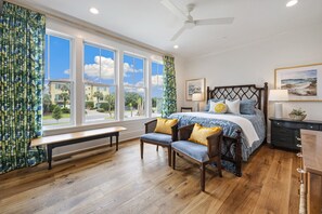 A bedroom with a large window, a bed with a headboard, two chairs, a bench, a ceiling fan, and artwork on the walls. - Cornfed Pelican 9 Bedroom Home (Georgetown)