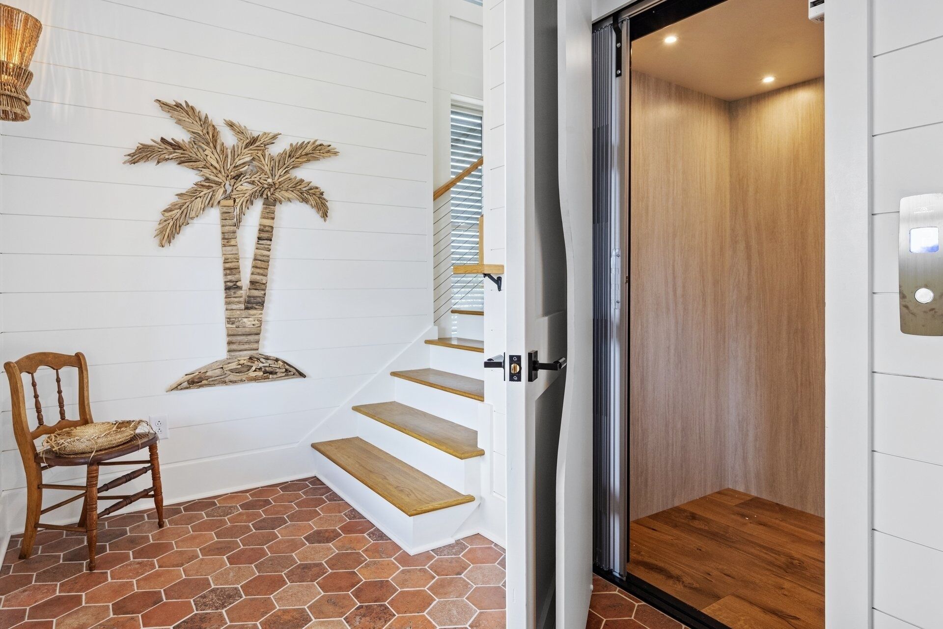 A staircase with wooden steps leading to an elevator, a chair with a woven seat, and a wall with a palm tree decoration.