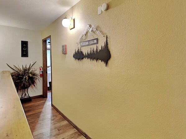A hallway with a wooden floor, a wall-mounted light fixture, and a decorative wall piece depicting mountains and trees.