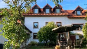 A two-story house with a red-tiled roof, a covered patio with chairs, and a small garden area.