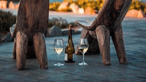 Dos sillas de madera con una botella de vino y dos copas en una terraza de madera frente al mar.