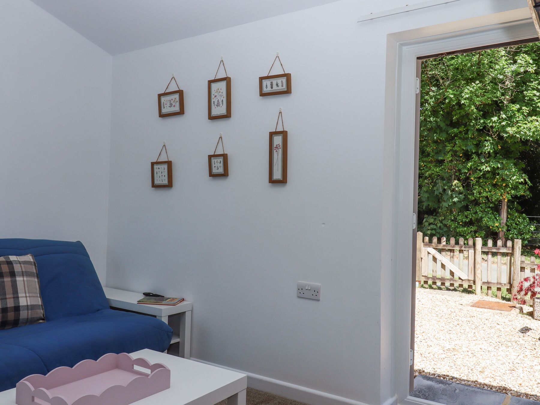 Una habitación con un sofá azul, una mesita de noche blanca con un libro y una mesa de centro blanca. Seis cuadros enmarcados adornan la pared, y se puede ver una valla de madera a través de la puerta.