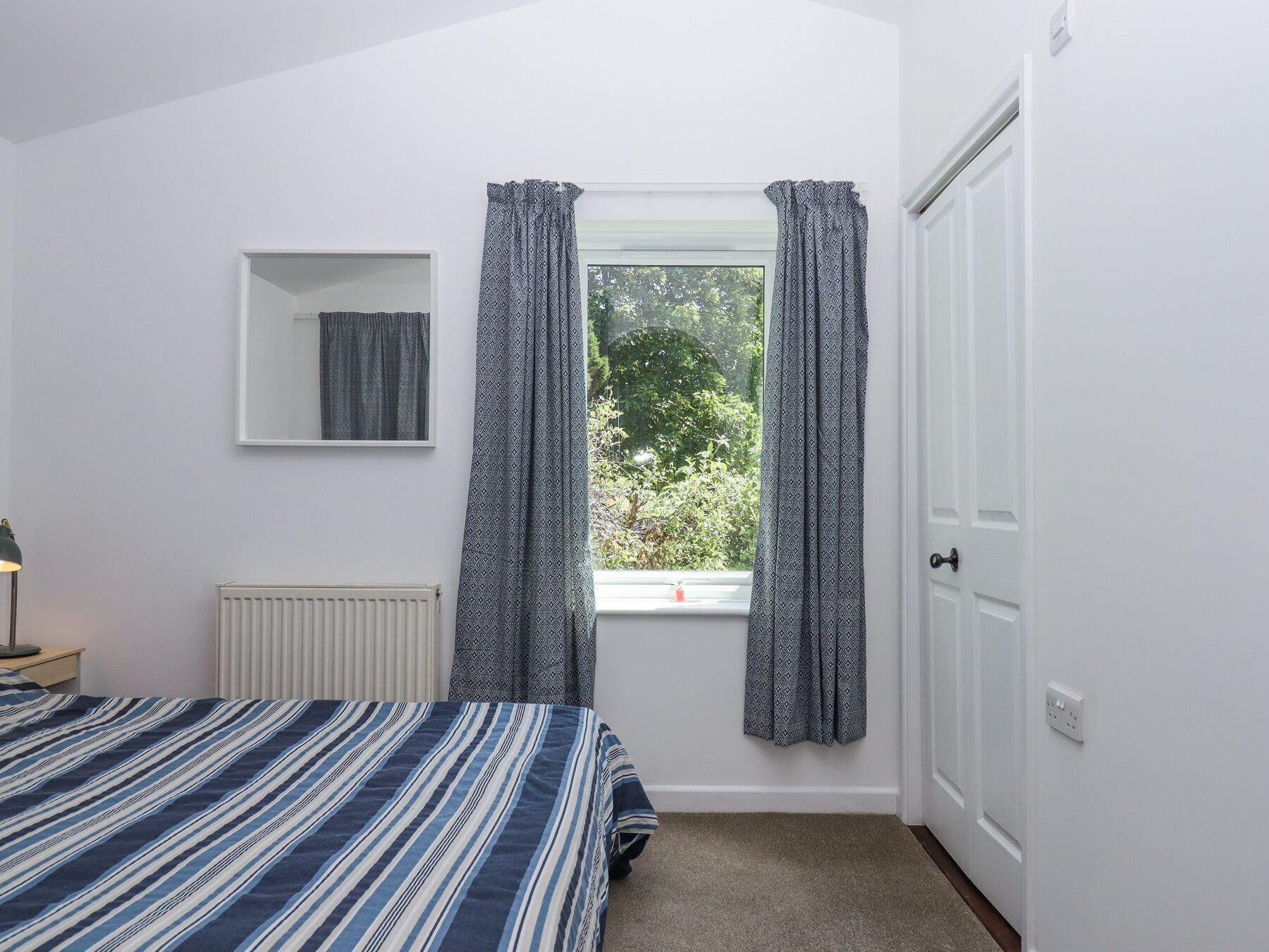 A bedroom with a bed, a radiator, a window with patterned curtains, a mirror, and a door.