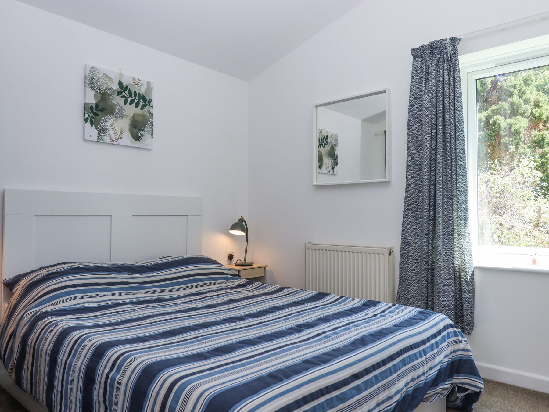 A bedroom with a bed, a bedside table with a lamp, a radiator, a window with curtains, and a wall-mounted mirror.