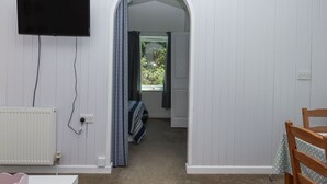 Una habitación con pared blanca, un televisor de pantalla plana montado sobre un radiador blanco, una silla de madera con mantel de lunares y una ventana con cortinas.