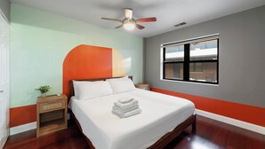 A neatly made bed with white linens, a wooden nightstand with a small plant, a ceiling fan, and a window with a view of a building. - Cozy Condo, Sleeps 2! (Chicago)