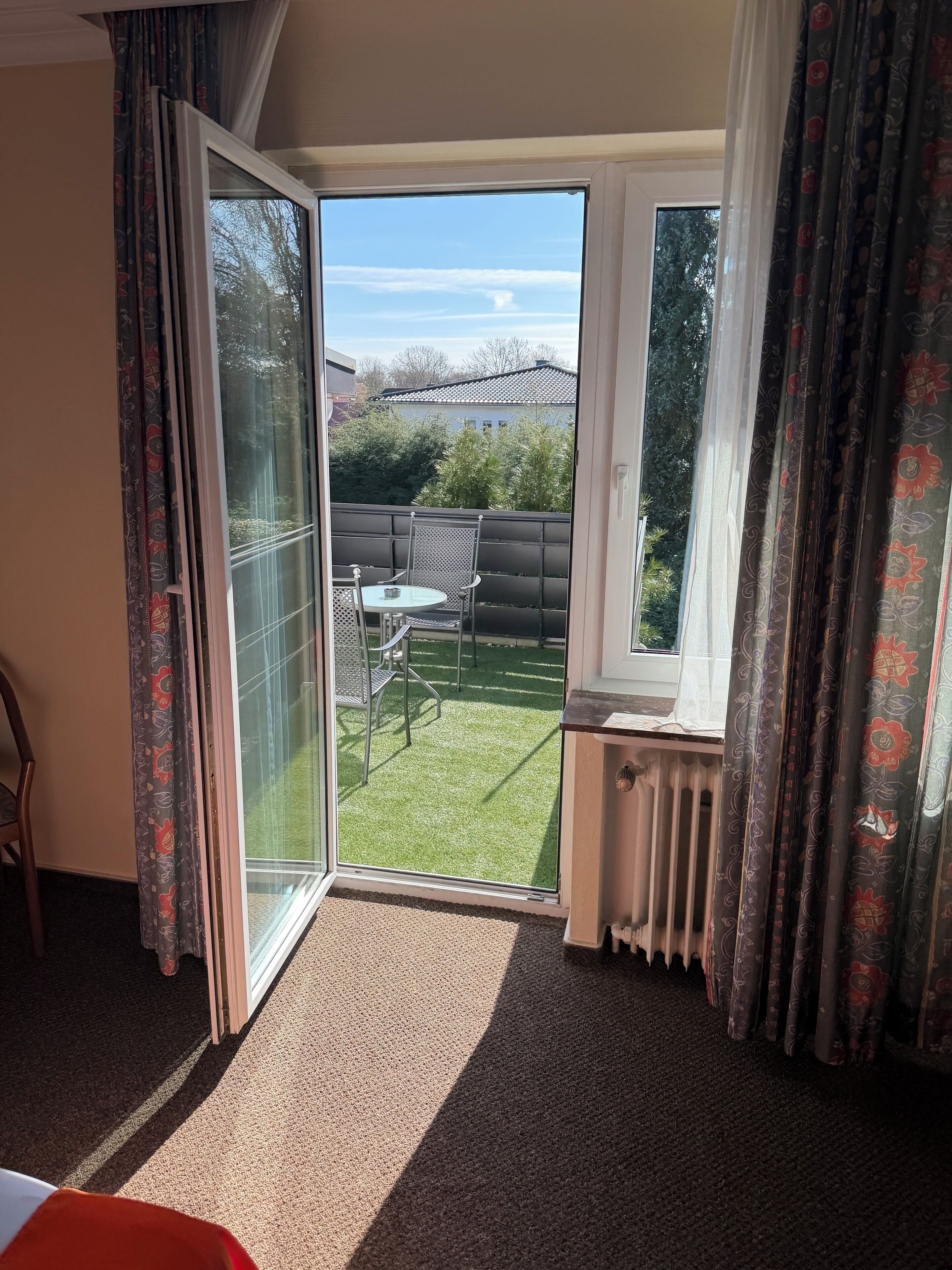 Ein Zimmer mit einer Schiebeglastür, die auf einen Balkon mit Tisch und Stühlen führt, einem Heizkörper und gemusterten Vorhängen.