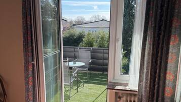 Ein Zimmer mit einer Schiebeglastür, die auf einen Balkon mit Tisch und Stühlen führt, einem Heizkörper und gemusterten Vorhängen.