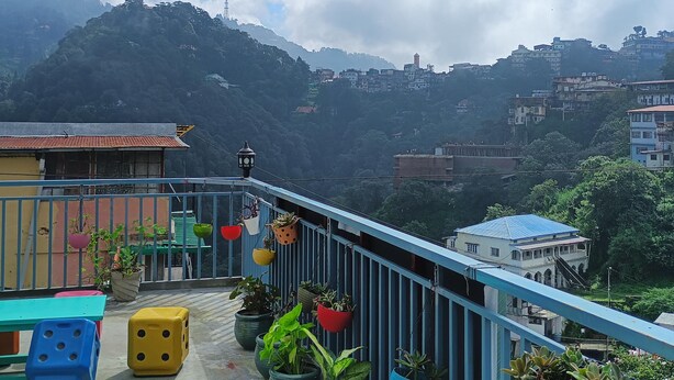 Ein Balkon mit Blumentöpfen, Blick auf eine gebirgige Landschaft und ein ferner Turm.