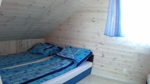 Een kleine kamer met een enkel bed, houten plafond en muren, een raam met gordijn, en een bed met een blauw beddegoed met patroon.