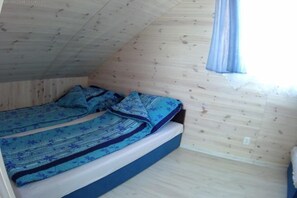 A small, single-bed room with a wooden ceiling and walls, a window with a curtain, and a bed with a blue patterned bedspread.