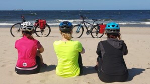 Drie personen zitten op een zandstrand, met fietsen en helmen, en kijken uit over de oceaan.