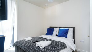 Ein ordentlich bezogenes Bett mit einer grauen, gemusterten Tagesdecke, weißen und blauen Kissen sowie einem aufgerollten weißen Handtuch.