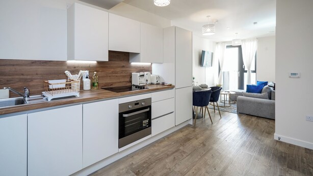 Eine moderne Küche mit weißen Schränken, einer Holz Arbeitsplatte und einem eingebauten Ofen. Es gibt einen Essbereich mit einem blauen Stuhl und einem grauen Sofa.