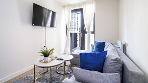 Ein modernes Wohnzimmer mit einem grauen Sofa, blauen Kissen, einem kleinen runden Tisch mit einer Pflanze und einem an der Wand montierten Fernseher.