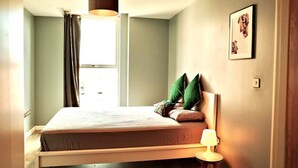 A bedroom with a bed, a bedside table with a lamp, a window with curtains, and a wall-mounted picture. - Central 2BR Birmingham Apt - Free Parking (Birmingham)