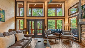 A spacious living room with a brown leather sofa, a wooden coffee table, and a stone fireplace. Large windows offer views of the surrounding forest. - Olivers Modern Twist- Cherry Log GA (Cherry Log)