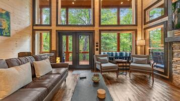 A spacious living room with a brown leather sofa, a wooden coffee table, and a stone fireplace. Large windows offer views of the surrounding forest.