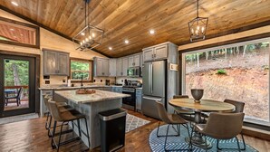 A modern kitchen with wooden ceiling, built-in cabinets, a central island, and a dining area with a view of the forest. - Olivers Modern Twist- Cherry Log GA (Cherry Log)
