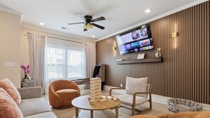 Ein modernes Wohnzimmer mit einem flachbildfernseher an der Wand, einem Deckenventilator und einer gemütlichen Sitzecke mit Sofas und Sesseln.