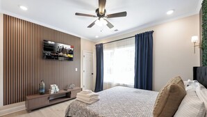 Ein modernes Schlafzimmer mit Bett, Deckenventilator, montiertem Fernseher und einem Fenster mit Vorhängen.