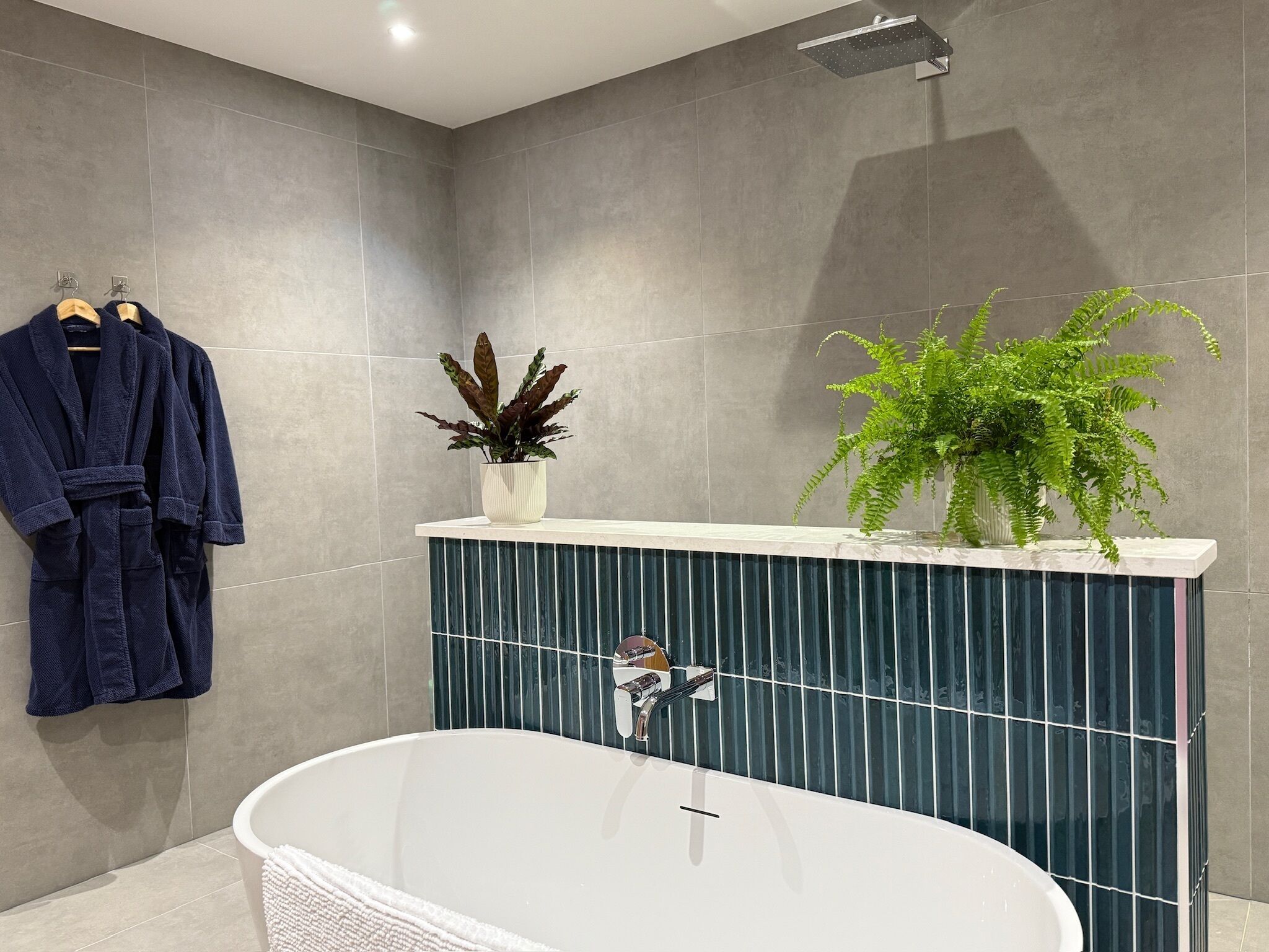 A modern bathroom with a freestanding bathtub, grey tiled walls, a towel rack with robes, and two potted plants on a vanity.