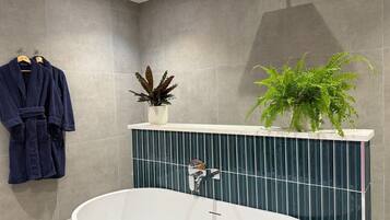 A modern bathroom with a freestanding bathtub, grey tiled walls, a towel rack with robes, and two potted plants on a vanity.