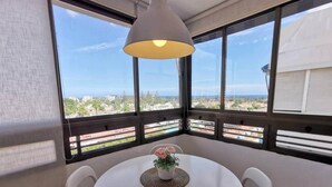 Una habitación con una mesa redonda, sillas blancas, una planta en maceta y un amplio ventanal con vistas a edificios y follaje.