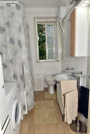 A compact bathroom with a washing machine, toilet, sink, and a window with a view of greenery. - Apartment Ljubica Dubrovnik Center (Dubrovnik)