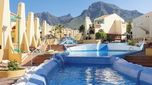 A swimming pool area with steps leading into the water, surrounded by buildings with balconies and a mountain in the background. - Home2Book Stylish Apt Fañabé, BBQ Terrace & Pool (Adeje)