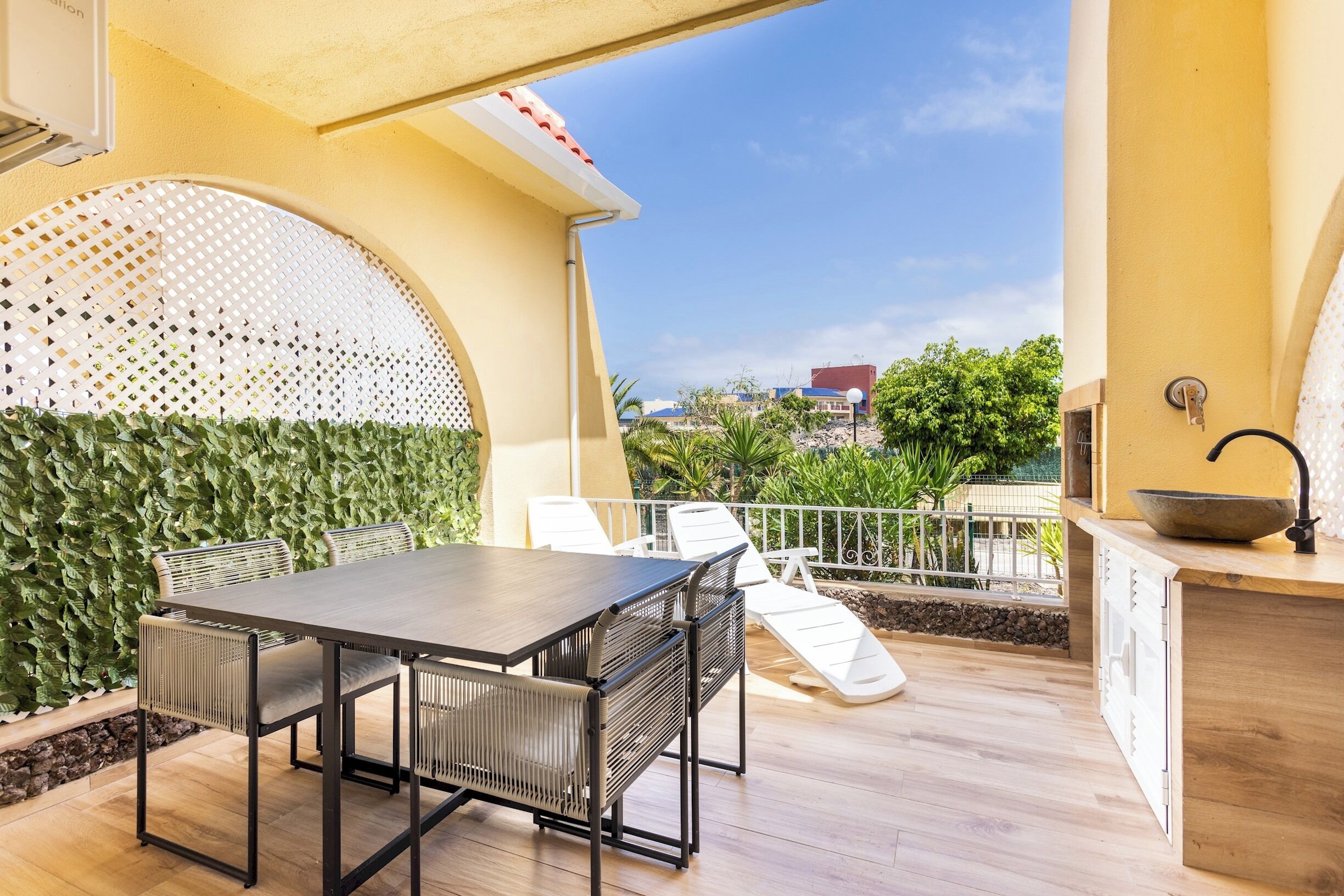 An outdoor patio with a dining table and chairs, a small kitchen area with a sink, and a view of greenery and buildings in the background.